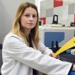 Zdjęcie portretowe: dr inż. Anna Masek w białym fartuchu siedzi prawym profilem przy biurku. W ręku trzyma żółte paski. Po prawej stronie ekran komputera z danymi. W tle laboratorium. 