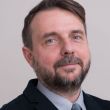 Portrait photo: Prof. Marcin Kamiński wearing a dark jacket, blue shirt and tie a against a grey background.