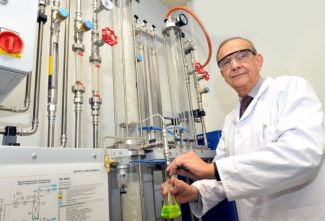 Zdjęcie portretowe: Prof. Andrzej Górak w białym fartuchu i okularach ochronnych stoi bokiem przy specjalistycznym urządzeniu.