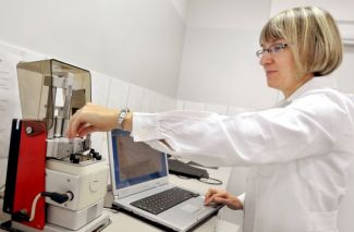 Zdjęcie portretowe: dr inż. Janina Leks-Stępień lewym profilem stoi w białym fartuchu przy specjalistycznym urządzeniu i komputerze i dokonuje pomiarów.