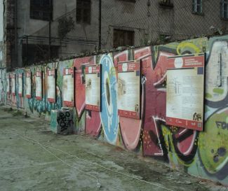 Mur w brzydkimi grafikami. W tle zniszczona zabudowa.