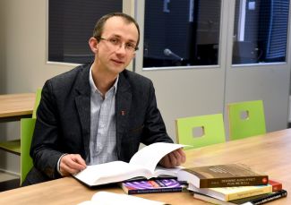 Zdjęcie portretowe: dr inż. Bartłomiej Stasiak siedzi przy stole, na którym leży stos książek. Jedna z nich otworzona. Na drugim planie zielone krzesła i niebieskie szyby kabiny.