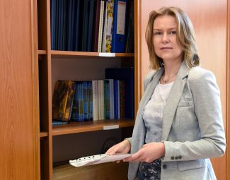 Zdjęcie portretowe: dr inż. Edyta Marcinkiewicz stoi przy szafie z półkami z segregatorami i książkami.