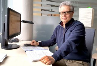 Portrait photo: Dr Eng. Jakub Szczepaniak is sitting sideways at a white desk. On the tabletop an open thick notebook and a computer screen.
