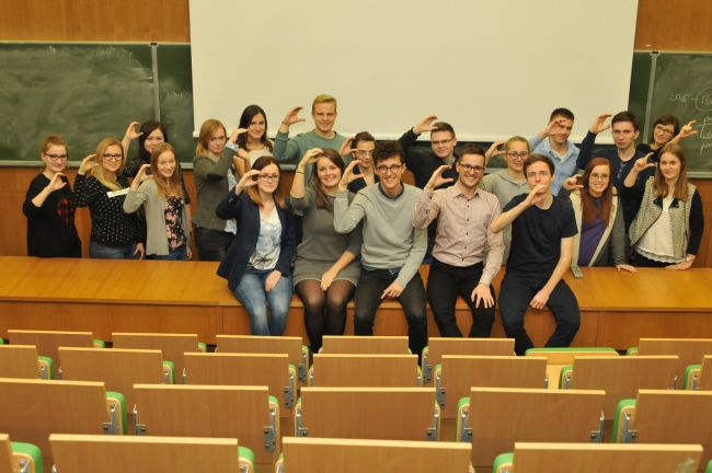 Portrait photo: a group of about 30 students from the Student Science Club Cirkula in a lecture theatre on the stage. Their right hands are arranged in the letter C. At the back there is a whiteboard and some green school boards.
