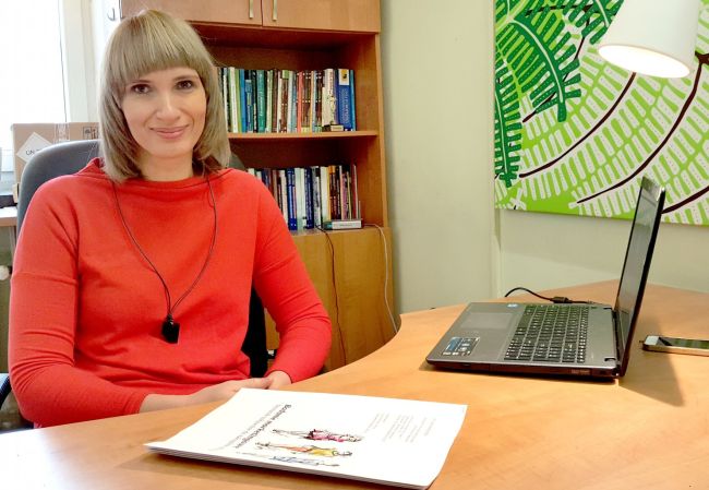 Zdjęcie portretowa: dr hab. Małgorzata Koszewska w czerwonej bluzce siedzi przy biurku. NA nim dokumenty i komputer. W tle zielona grafika i regał z książkami.