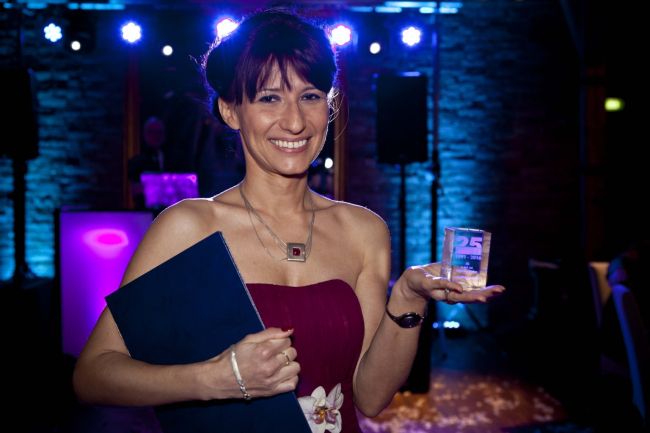 Portrait photo: Assoc. Prof. Eng. Magdalena Grębosz-Krawczyk, TUL Prof. in a ball gown, is presenting a statuette and a diploma. Backlit stage in the background.