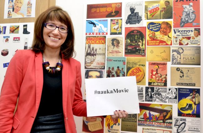 Portrait photo: smiling Assoc. Prof. Eng. Magdalena Grębosz-Krawczyk, TUL Prof. wearing a red jacket, is holding a white card with the inscription Science Movie. Colourful pictures in the background.