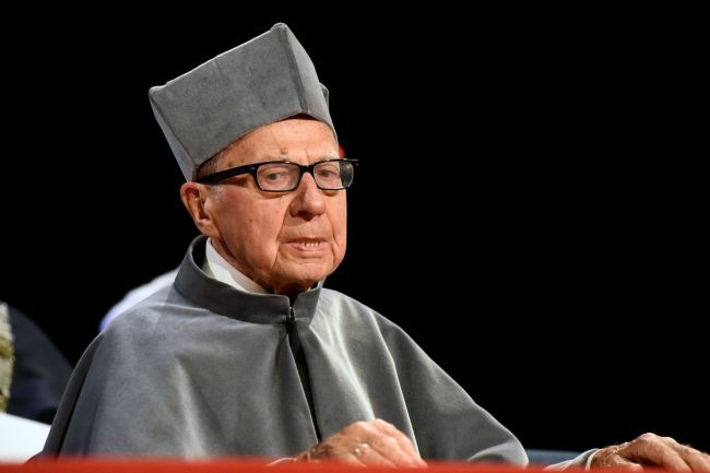 prof. Edward Kącki w szarej todze i birecie siedzi na tle czarnej ściany.