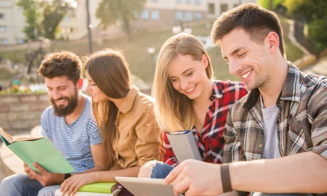 Czworo uśmiechniętych studentów (dwie studentki w środku) przeglądają materiały informacyjne. W tle park.