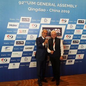 Elegancko ubrany prof. J. Wojewoda z prezydentem UIM- Rafelle Chiulli na tle niebieskiej ścianki reklamowej z logami firm.