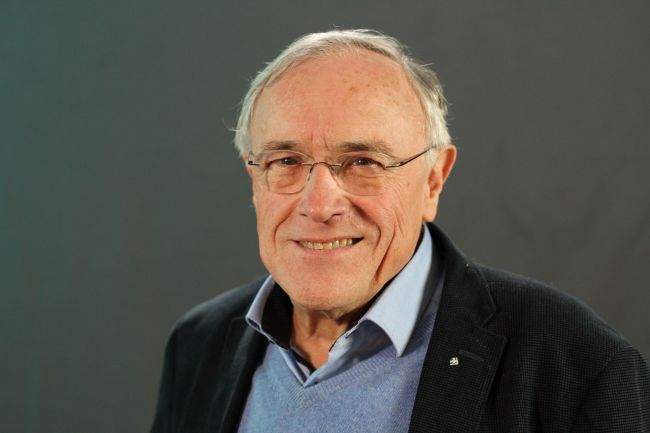 Zdjęcie portretowe: prof. Klaus Müllen w ciemnej marynarce i niebieskiej koszuli na szarym tle.