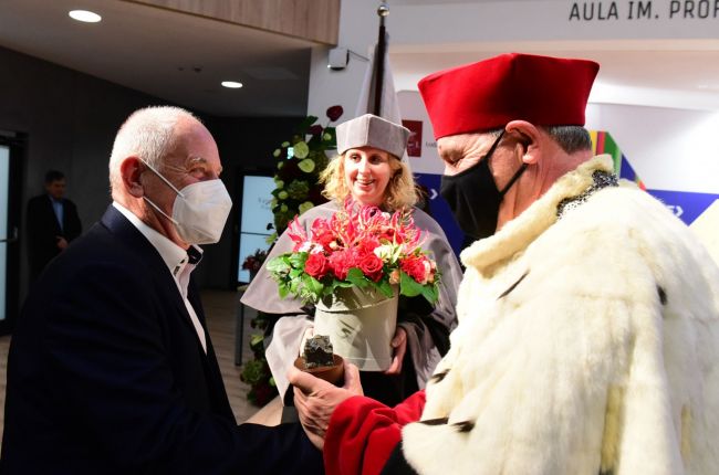 Bokiem stoją: po prawej stronie Rektor PŁ w todze i gronostajach, po lewej stronie dyr. Syriusz-Wolski w garniturze. W tle dyr. D. Piotrowska w todze trzyma bukiet kwiatów.