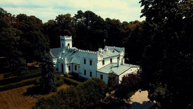 Rozłożysty, biały dworek o nieregularnym kształcie wśród wysokich drzew.