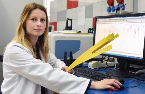 Zdjęcie portretowe: dr inż. Anna Masek w białym fartuchu siedzi prawym profilem przy biurku. W ręku trzyma żółte paski. Po prawej stronie ekran komputera z danymi. W tle laboratorium. 