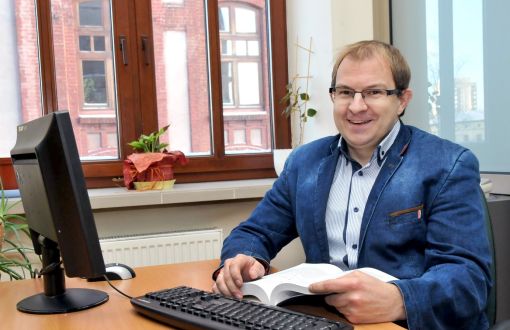 Zdjęcie portretowe: prof. Grzegorz Szymański w niebieskiej marynarce siedzi przy biurku i komputerze.