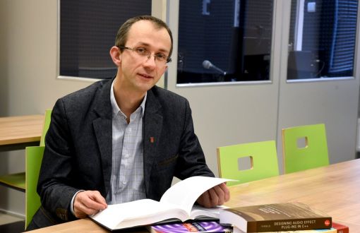 Zdjęcie portretowe: dr inż. Bartłomiej Stasiak siedzi przy stole, na którym leży stos książek. Jedna z nich otworzona. Na drugim planie zielone krzesła i niebieskie szyby kabiny. 