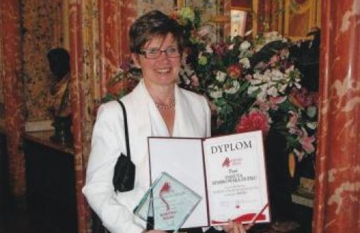 Danuta Kembłowska-Dupieu w białej marynarce trzyma w dłoniach i prezentuje dyplom i statuetkę. W tle elegancie wnętrze z kwiatami.