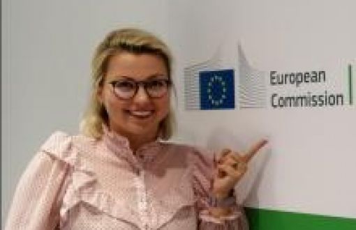 Zdjęcie portretowe: uśmiechnięta Kaja Kantorska na tle jasnej ściany z logiem European Comission, które wskazuje palcem wskazującym.