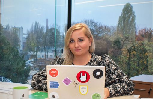 Sylwia Kobus, absolwentka PŁ, przy biurku i laptopie. W tle przeszklone okno z widokiem na park.