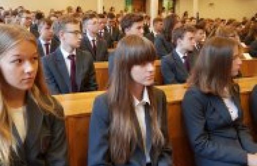 Uczniowie Liceum Politechniki Łódzkiej w szarych marynarkach i bordowych apaszkach (uczennice) i krawatach (uczniowie) siedzą w auli przy drewnianych ławach.