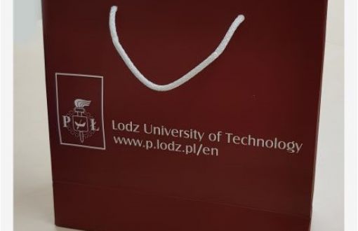 Bordowa, kwadratowa torba papierowa z białym uchem i napisem Politechnika Łódzka w wersji polskiej i angielskiej.