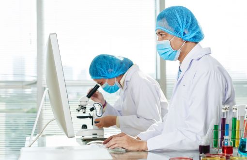 Laboratorium chemiczne. Dwoje badaczy w białych fartuchach i niebieskich czepkach i maseczkach w laboratorium stoją przy mikroskopie i ekranie.