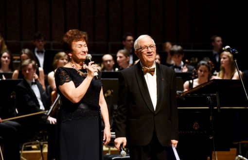 prof. Jan Krysiński w smokingu i Grażyna Sikorska w czarnej sukni na scenie Filharmonii Łódzkiej. W tle orkiestra.