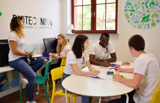 Pięciu studentach w białych koszulkach rozmawia w sali zajęć.