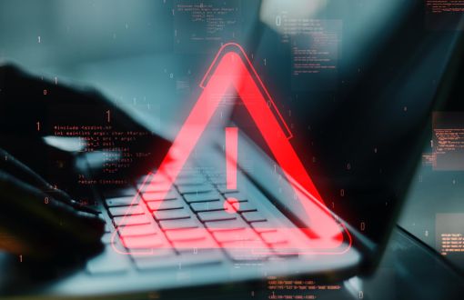 grafika przedstawiająca palce piszące po klawiaturze laptopa, a na nich hologram trójkątaczerwonego z wykrzyknikiem wewnątrz
