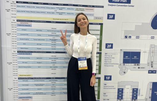 młoda uśmiechnięta dziewczyna w biało-czarnym stroju stoi na tle ścianki z logo konferencji ISMAR